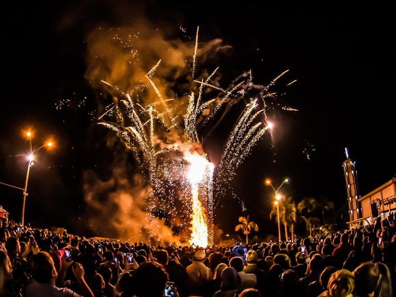 Fogueira de São João queimando enquanto fogos de artifício explodem ao fundo e dezenas de pessoas assistem ao show