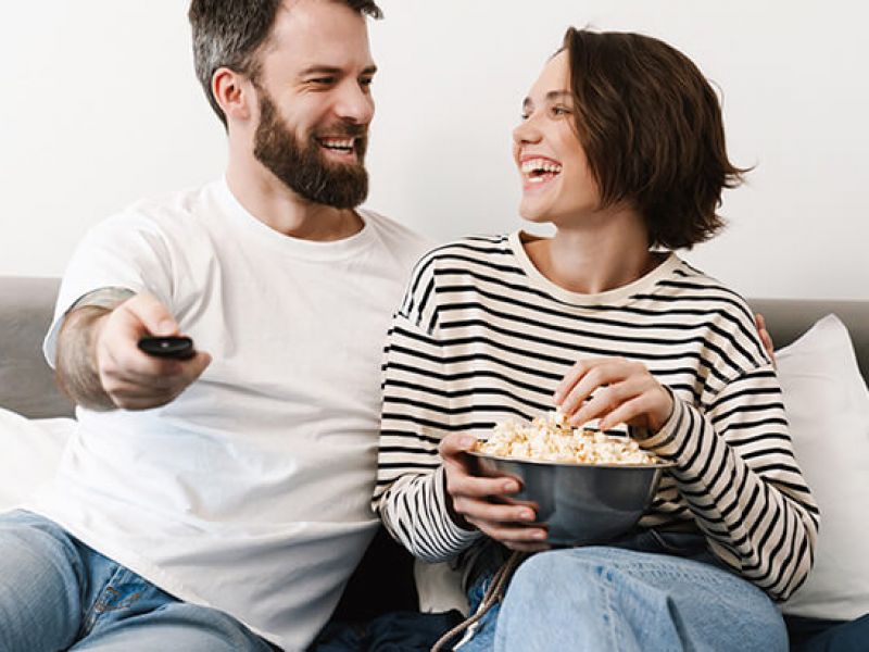 casal sorrindo segurando pipoca e controle de tv enquanto assistem séries sobre viagens