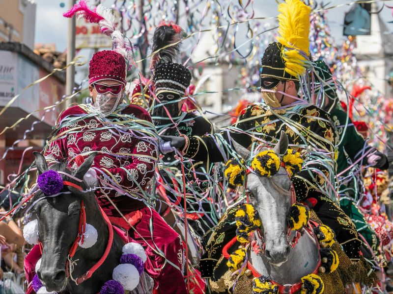 Quatro pessoas montadas a cavalo, fantasiadas com os trajes típicos e coloridos das Cavalhadas.