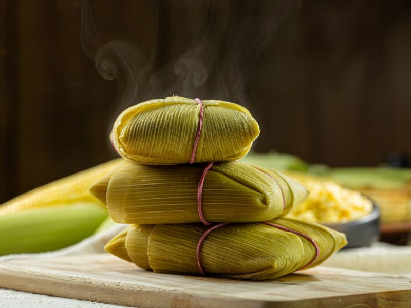 Três pamonhas embrulhadas na folha do milho, sobre uma táboa de madeira