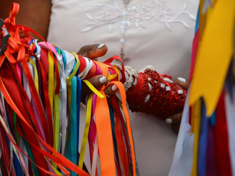 Parte dos braços e mãos de uma pessoa que seguram um dos bastões decorados e coloridos utilizados na Festa do Divino