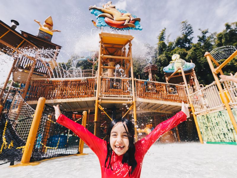 Criança de molhada, de braços abertos e sorrindo em frente ao parque aquático infantil do Hot Park