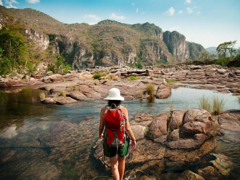 Pessoa de costas caminhando em um pequeno rio com pedras, e a Chapada dos Veadeiros ao fundo